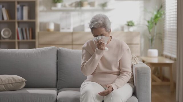 Senior woman sitting on home sofa blowing nose with tissue feeling sick miserable unwell and uncomfortable fighting severe cold flu virus infection during winter seasonal allergy in living room