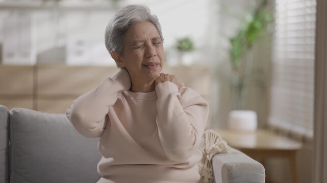Senior woman sitting on home couch inside living room experiencing severe neck pain shoulder ache stiff muscle tension and deep physical discomfort showing distressed emotion indoors