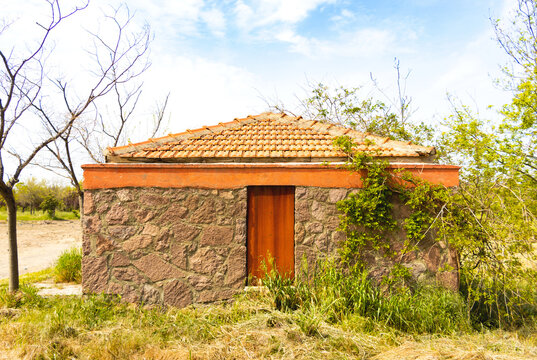 Rustic rural stone outbuilding featuring a weathered orange terracotta tile roof and a solid rusted iron door amidst wild overgrown grass.