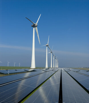 Aerial view of a row of wind turbines standing over a vast field of solar panels under a clear blue sky in Netherlands.