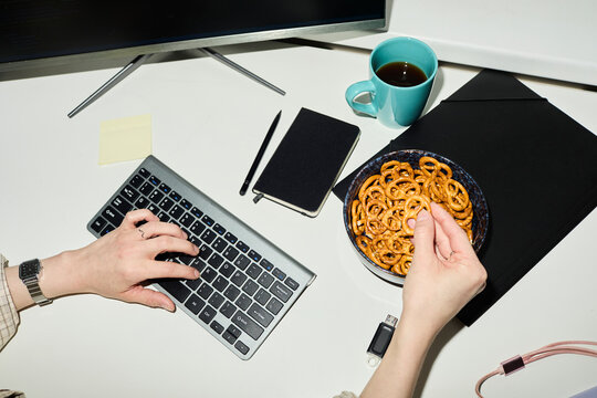 Employee typing at office table and eating pretzel on lunch break