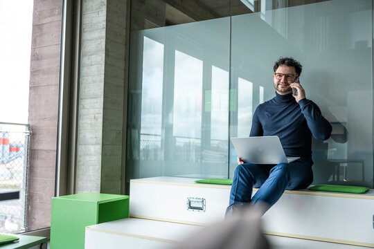 Smiling businessman with laptop talking on mobile in office