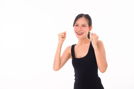 A happy young woman in a black tank top smiles brightly with fists raised, expressing excitement and confidence against a plain white background.