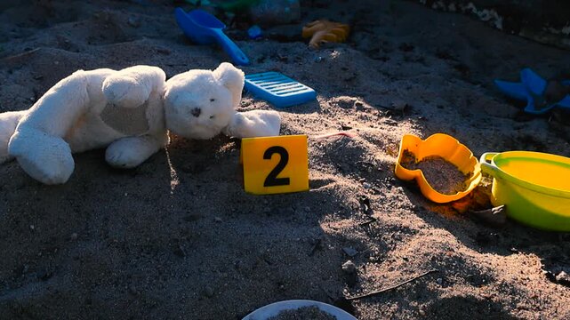 Abandoned toy bunny and colorful plastic toys lying in a sandbox next to a yellow crime scene evidence marker, conceptualizing the loss of innocence or a child endangerment investigation