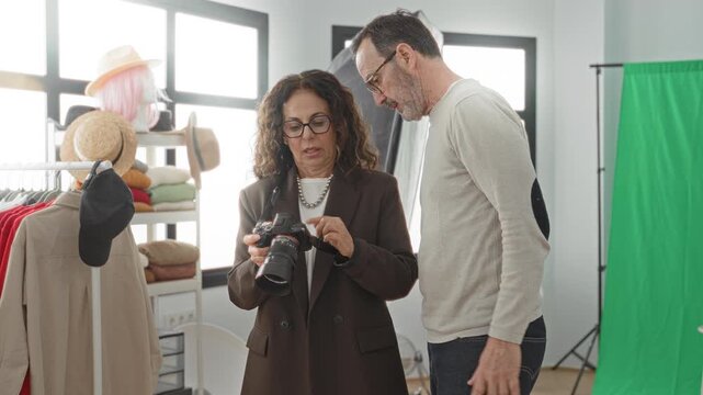 Middle aged woman holds camera and reviews settings while a man leans in beside a clothing rack and green screen in a studio; collaboration.