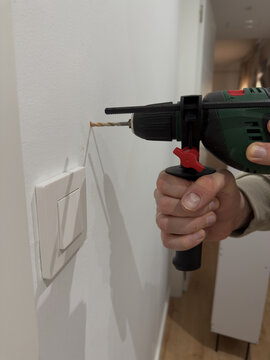 Male hand drills hole in white wall near light switch using green corded electric drill with white shoe cabinet visible in hallway background