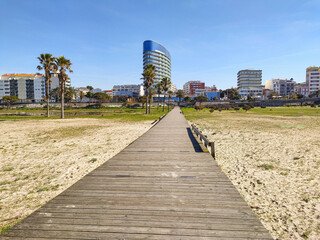 Fototapeta premium Discover a path to modern tranquility. This wooden boardwalk offers a stunning perspective towards a coastal city with iconic architecture and palm trees under a vibrant blue sky.