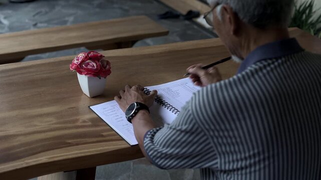 Over the shoulder shot of old Asian man writing note on book with blurred foreground and head at wooden table