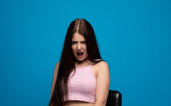 Young woman shouting and expressing frustration, anger, and aggression with mouth open on blue background