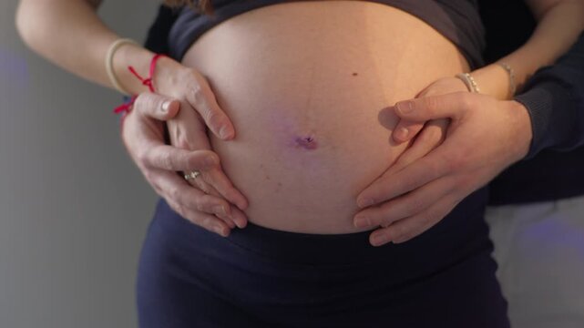 Close view of pregnant belly with warm side lighting, both hands placed gently from either side, bracelet visible on one wrist
