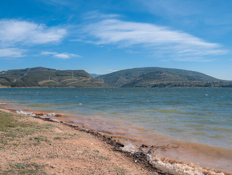 A very windy day with quite strong waves reaching the shore of the B&aacute;rcena del Bierzo reservoir, the water is choppy; in the background, mountains with electricity pylons and a blue sky can be seen