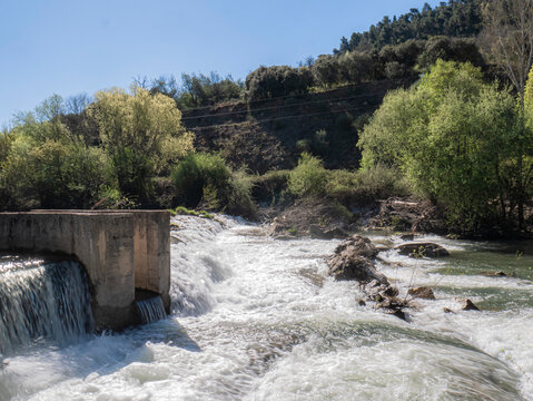 A long-exposure image of a fast-flowing river cascading down a small dam amid lush vegetation and wooded hills, under a clear, bright sky