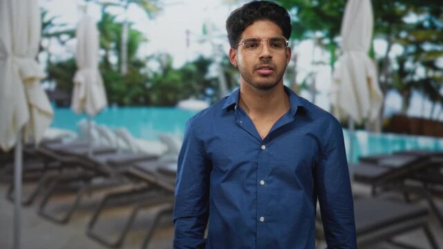 Man in blue shirt and glasses rubs neck by poolside lounge chairs and umbrellas near palm trees in building; contemplative vacation.