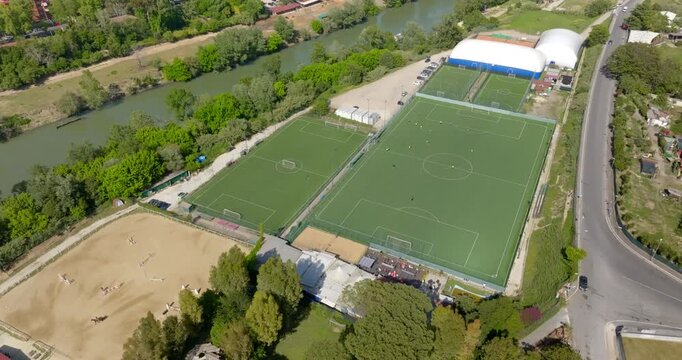 High-angle aerial view of a modern sports center featuring multiple green synthetic grass soccer fields, located in a scenic outdoor area near a river and lush trees under bright daylight.
