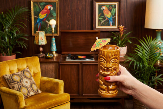 Hand holding a tiki cocktail mug with an umbrella in a retro 70s living room. Vintage interior features wood paneling, yellow armchair, and parrot art.