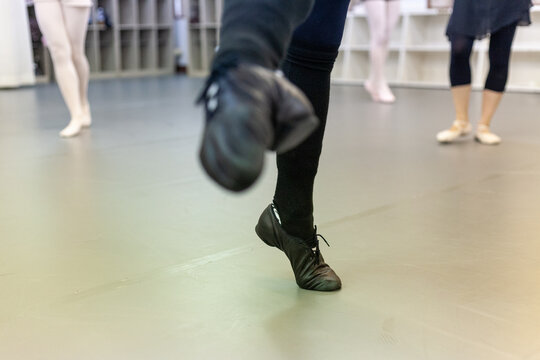 Dance class in a studio, close-up on a dancer's feet in black jazz shoes in motion, with ballet dancers wearing pointe shoes in the background.
