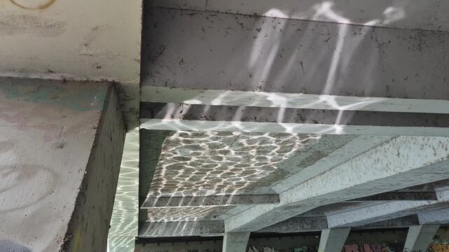 Sunlight reflects off water, creating shimmering caustic patterns on the concrete underside of a bridge. The mesmerizing light dance shifts gracefully across the urban structure.