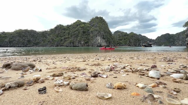 Sandy beach covered in seashells with limestone karst mountains and kayakers in Halong Bay Vietnam background
