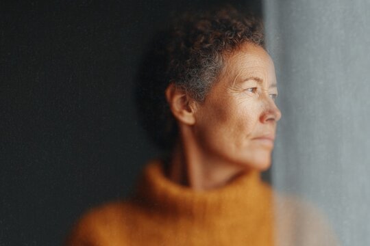 A woman in an orange turtle neck sweater leaning against a wall. she has a warm and inviting expression on her face and her hair is pulled back in a neat bun.