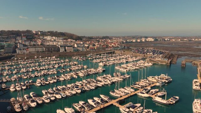 St Peter Port Harbour Guernsey.Low forward drone footage over QEII Marina and boats on moorings with town and Belle Greve Bay behind at low tide in late afternoon sun with calm clear blue water