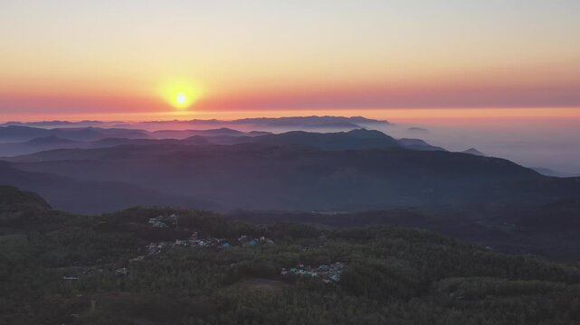 Yercaud's forested hills emerge from morning fog as the sun rises over layered mountain ridges in Salem Tamil Nadu, India. Slow wide angle drone shot captures the atmospheric valley and village below.