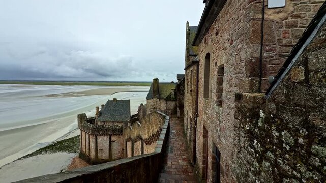 Stone walls of Mont-Saint-Michel Abbey stretch towards expansive tidal flats under a cloudy sky. Perfect for historical documentaries or travel features.