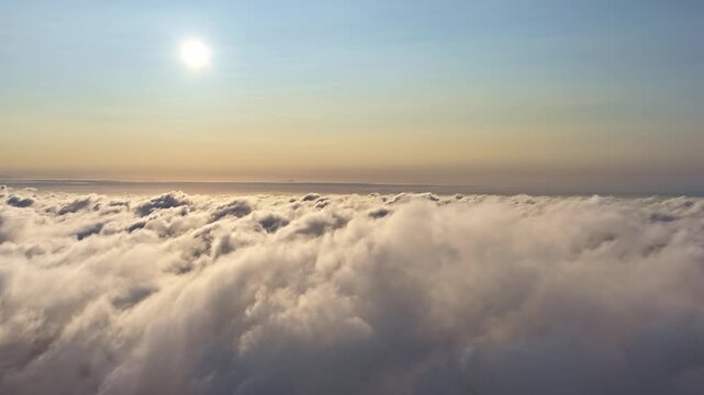 Breathtaking aerial pullback view flying above thick white cloud layer with glowing sun near warm golden horizon and deep blue sky above