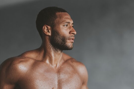 A shirtless man standing in front of a gray background looking away from the camera with a determined expression on his face as if he is ready to take on any challenge that comes his way.