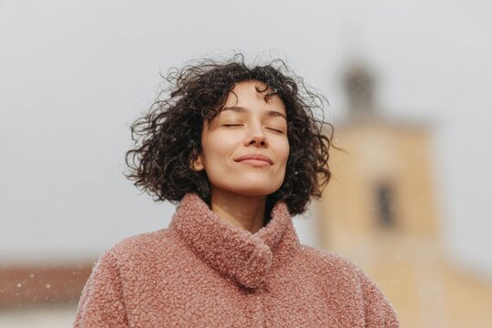 A woman with curly hair wearing a pink turtleneck sweater standing in front of a building with a blurred background.