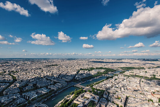 Aerial view of the Seine river winding through the historic urban landscape and dense architecture under a bright blue sky with scattered clouds in Paris, Ile-de-France, France.