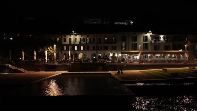 Illuminated waterfront promenade and buildings of Malgrate on shore of Lake Como at night in Italy. Aerial lateral view