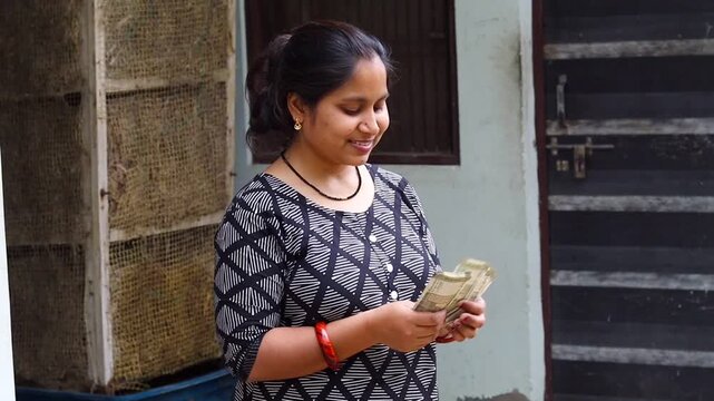 Smiling Indian woman counting five hundred rupee notes standing outside her house, happy female holding cash currency, authentic middle class household finance and savings concept
