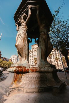 Low angle view of a vintage stone caryatid fountain on a Barcelona street with historic city buildings and palm trees in the background.
