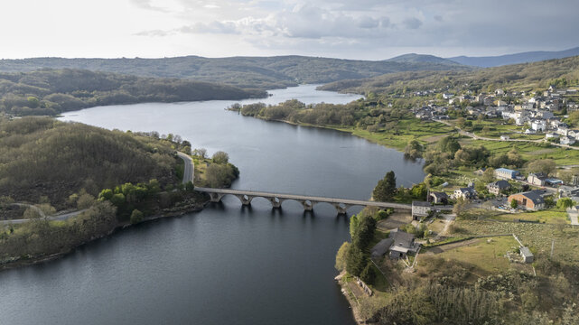 Aerial view of Encoro de Prada reservoir and a stone arch bridge crossing the water, surrounded by lush green hills and the village of A Veiga, Ourense, Spain.