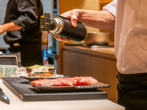Chef and team using blowtorch to sear fresh sashimi on plate in Japanese restaurant kitchen, close up of hand holding culinary torch flame, gourmet seafood preparation, fine dining sushi restaurant