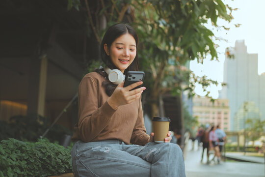 Young Asian woman smiling while using a smartphone and holding a coffee cup in an urban city park. Modern lifestyle, mobile connectivity, and wellness during a summer break in a natural outdoor