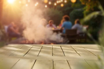 Fototapeten Fallen Empty wooden table with clean copy space for product placement in the foreground. Blurred background with garden camping scene with barbecue grill and people enjoying outdoor time, warm, relaxed.  © magdal3na