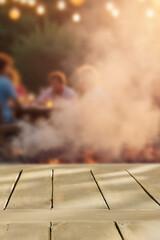 Fototapeta premium Empty wooden table with clean copy space for product placement in the foreground. Blurred background with garden camping scene with barbecue grill and people enjoying outdoor time, warm, relaxed.
