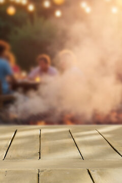 Empty wooden table with clean copy space for product placement in the foreground. Blurred background with garden camping scene with barbecue grill and people enjoying outdoor time, warm, relaxed.