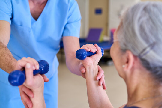 Senior Woman Doing Arm Exercises With Blue Dumbbells During Physiotherapy Session With Male Therapist In Rehab Center Indoors