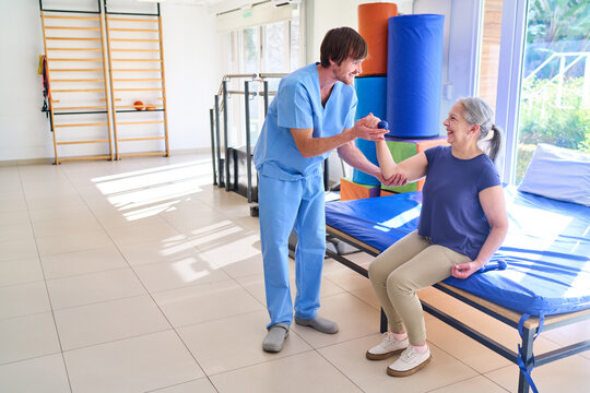 Senior Woman Lifting Dumbbell With Physiotherapist Assistance During Rehabilitation Session Indoors At Rehab Center For Recovery And Strength