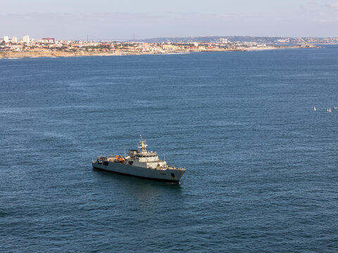 Aerial view of a grey naval patrol vessel sailing on the blue ocean with the coastal skyline in the background Cascais, Portugal.