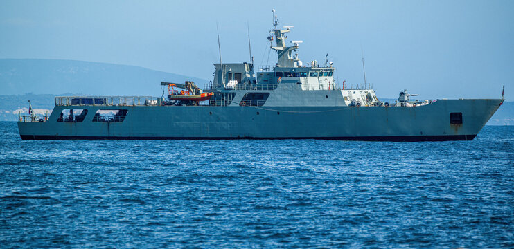 Aerial view of a grey naval patrol vessel sailing on the blue sea with a distant coastline under a clear sky in Cascais.