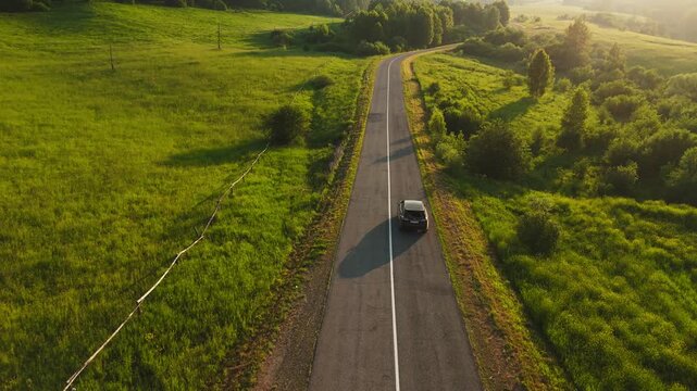 Aerial view of car driving down winding rural road. Golden sunlight bathes vibrant green fields. Road curves gently through peaceful countryside landscape. Car moves steadily forward