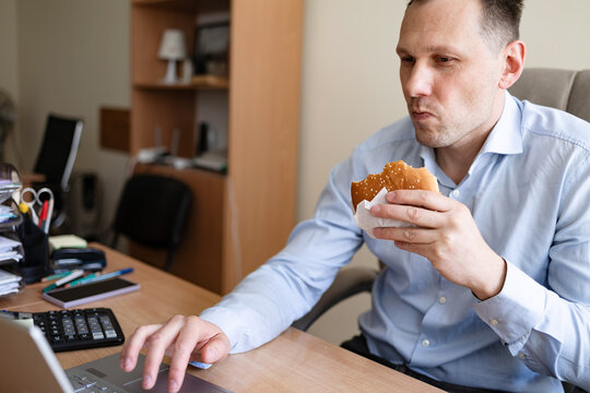 Caucasian male worker in office having fast food lunch at desk. Portrait of businessman eating burge
