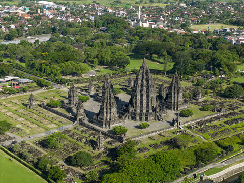 Aerial view of the ancient stone towers of Prambanan Temple surrounded by lush green parkland and the urban landscape of Prambanan, Daerah Istimewa Yogya, Indonesia.
