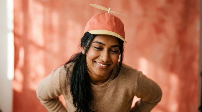 Warm indoor portrait of cheerful woman wearing coral propeller hat with cream blade, peach wall sunlight, relaxed smile, cozy knit sweater, playful lifestyle fashion and soft cinematic atmosphere