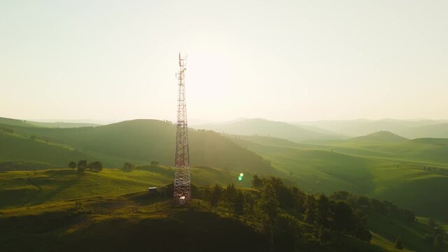 Aerial view of tall communication towers atop a grassy hill. Sunlight glows warmly over rolling green hills. Mist hovers softly in valley below. Landscape serene, untouched, and expansive