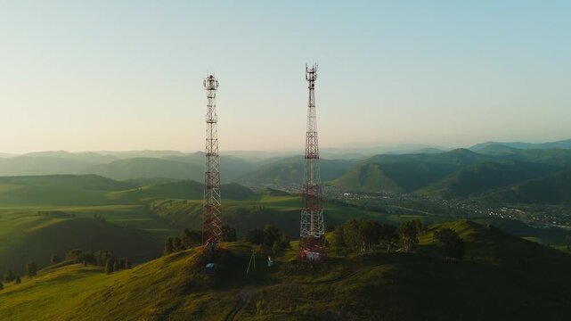 Two tall communication towers stand atop a grassy hill. Rolling green hills fade into distant mountains under clear skies. The scene is serene, untouched by human activity. Perfect for tech, travel