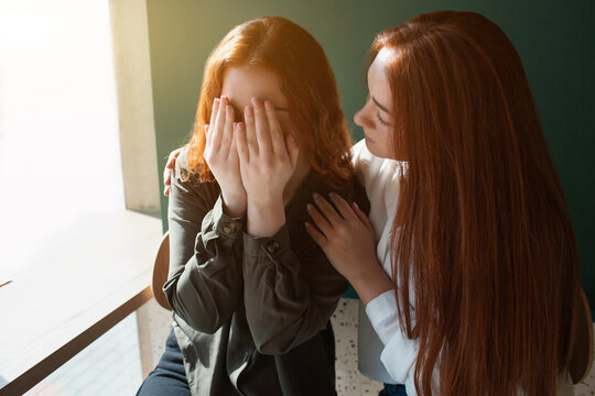 Young woman crying in a cafe covers her face with hands. Female model hugs her friend.